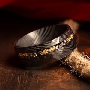 Puede incluir: Primer plano de un anillo negro con un patrón único y una incrustación dorada. El anillo está hecho de un metal oscuro con una superficie texturizada. La incrustación dorada está hecha de pequeños trozos de oro. El anillo está sobre una superficie marrón.