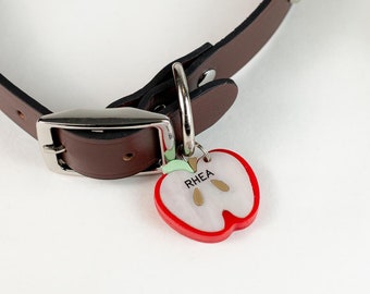May include: A brown leather dog collar with silver studs and a silver buckle. A red apple-shaped dog tag with the name "RHEA" is attached to the collar.
