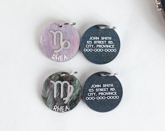 May include: Two round pet ID tags with a zodiac symbol and the name Rhea. The tags are on a white surface. One tag is a dark blue with a Virgo symbol and the other is a light purple with a Capricorn symbol. Both tags have the text "JOHN SMITH" and contact information.