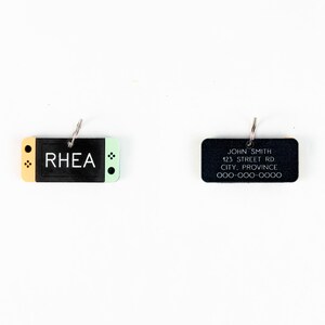 May include: Two black rectangular keychains with silver rings. The keychain on the left is shaped like a video game console with a yellow top and a green bottom. The text "RHEA" is printed in white on the black console. The keychain on the right has the text "JOHN SMITH 123 STREET RD CITY, PROVINCE 000-000-0000" printed in white.