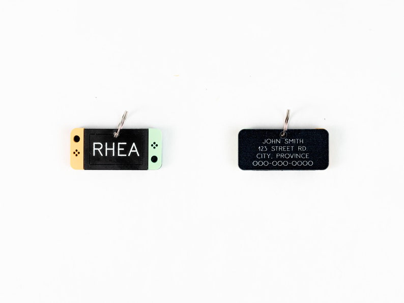 May include: Two black rectangular keychains with silver rings. The keychain on the left is shaped like a video game console with a yellow top and a green bottom. The text "RHEA" is printed in white on the black console. The keychain on the right has the text "JOHN SMITH 123 STREET RD CITY, PROVINCE 000-000-0000" printed in white.