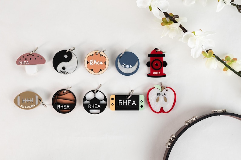 May include: Ten different personalized dog tags with various designs, including a mushroom, yin yang, flower, crescent moon, fire hydrant, basketball, soccer ball, video game controller, apple, and football. Each tag features the name "Rhea" and some have additional text.