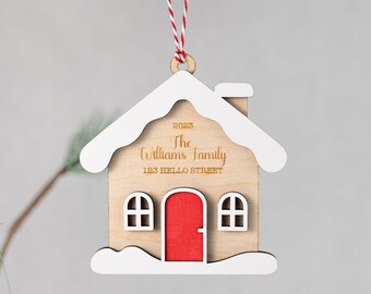 Puede incluir: Un adorno navideño de madera con forma de casa con una puerta roja y detalles de nieve blanca. El adorno está personalizado con el texto "2023 The Williams Family 123 HELLO STREET".