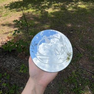 Peut inclure: Un miroir circulaire présentant un motif gravé représentant un visage de diable de dessin animé avec des oreilles pointues et des cornes. La surface réfléchissante montre le ciel et les nuages, tandis que le motif gravé est blanc mat.
