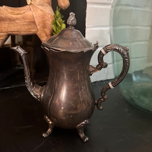 May include: Antique silver-plated teapot with a decorative handle and lid finial. The teapot has a curved spout and rests on three small feet. The handle features white accents. The teapot is displayed on a dark surface.