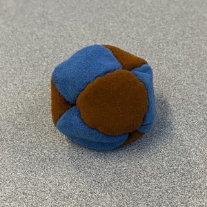 May include: A small, multi-sided fabric ball with a patchwork design. The ball features alternating blue and brown panels, creating a geometric pattern. The soft texture suggests a toy or decorative item.