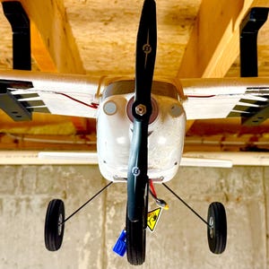 May include: A white and silver model airplane with a black propeller hanging from the ceiling. The airplane has a black and white striped tail and black landing gear.