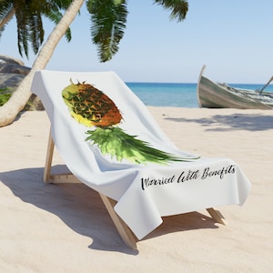 May include: White beach towel with a watercolor pineapple design and the text "Married With Benefits". The towel is draped over a wooden beach chair on a sandy beach with palm trees and the ocean in the background.