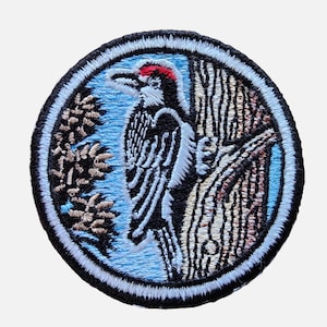 May include: Embroidered patch featuring a black and white woodpecker perched on a tree trunk. The bird has a red crest and is surrounded by blue and brown foliage.
