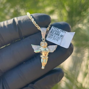 May include: A gold chain necklace with a diamond-encrusted angel pendant. The pendant is about 2 inches tall and features a cherub with wings outstretched. The tag attached to the chain reads "PEND MEN SJ258D $2,599.00 10K 0.13CT 203 2310".