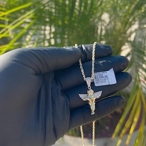 May include: A gold chain necklace with a diamond-encrusted angel pendant. The pendant is about 1 inch tall and features a cherub with wings outstretched. The chain is a rope chain style. The tag attached to the chain reads "PEND MEN SJ259D $2,599.00 10K 0.13CT 203'2310"