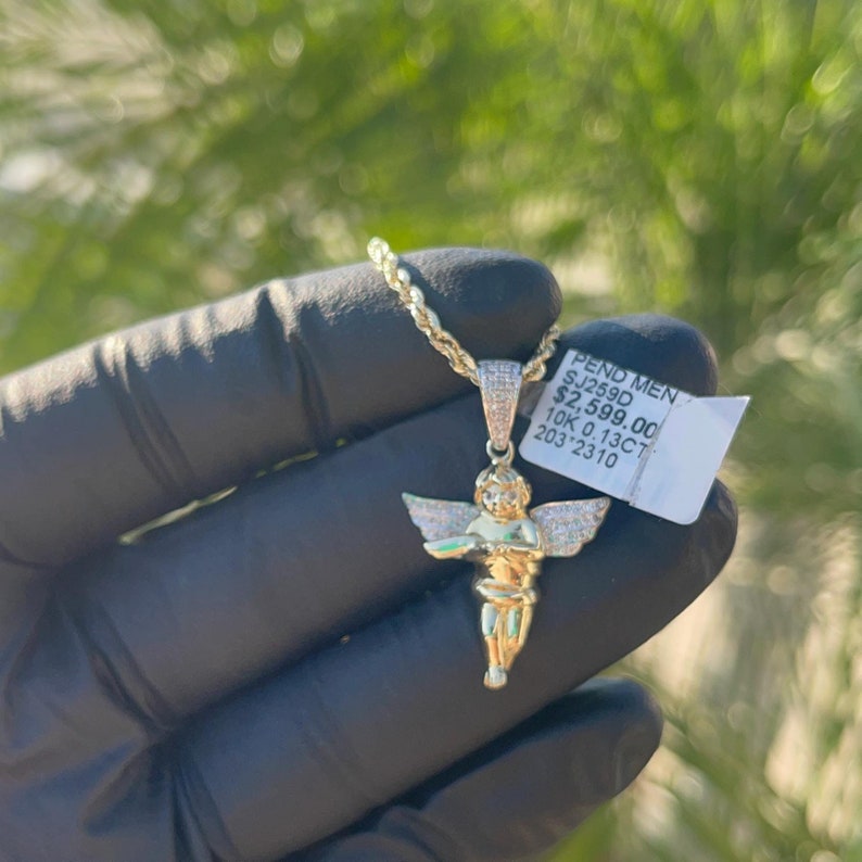 May include: A gold chain necklace with a diamond-encrusted angel pendant. The pendant is about 2 inches tall and features a cherub with wings outstretched. The tag attached to the chain reads "PEND MEN SJ258D $2,599.00 10K 0.13CT 203 2310".