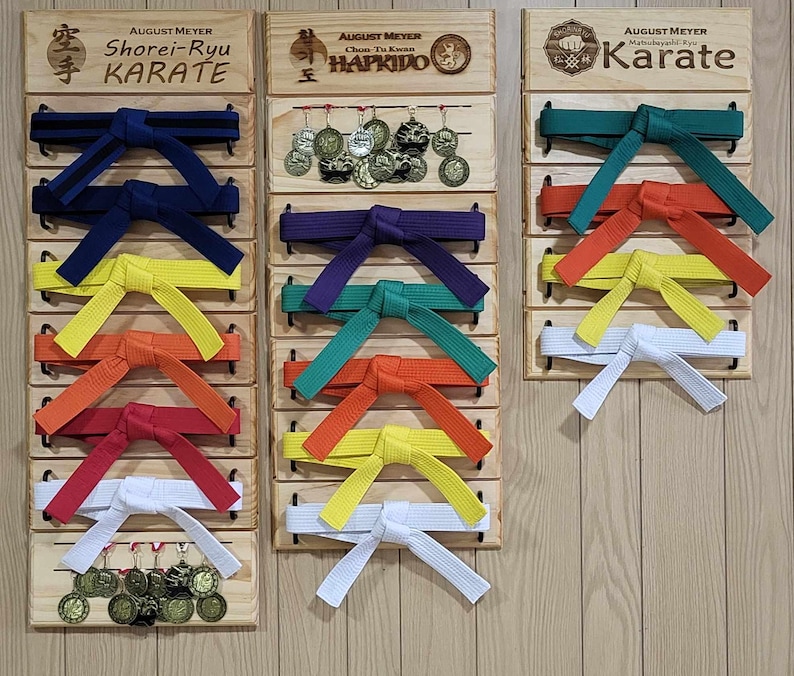 Op de afbeelding: Drie houten displayborden met rijen karategordels in verschillende kleuren, waaronder zwart, blauw, groen, geel, oranje, rood en wit. De borden zijn gelabeld met de namen van verschillende vechtsporten: Shorei-Ryu Karate, Hapkido en Karate.