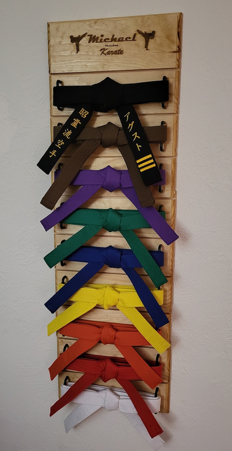 Op de afbeelding: Een houten karategordelrek met de naam "Michael Shito-ryu Karate" gegraveerd op de bovenkant. Het rek bevat een verscheidenheid aan karategordels, waaronder zwarte, bruine, paarse, groene, blauwe, gele, oranje, rode en witte gordels.