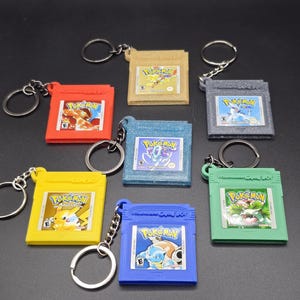 May include: A collection of keyrings, each with a miniature Game Boy cartridge in colours like red, yellow, and blue. Each cartridge has a Pokemon game label and a silver keyring. The cartridges feature various Pokemon characters.