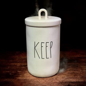 Puede incluir: Un bote de cerámica blanco con tapa y asa. La palabra "KEEP" está impresa en negro en la parte delantera. Sale vapor del bote, lo que sugiere que contiene una bebida o alimento caliente.