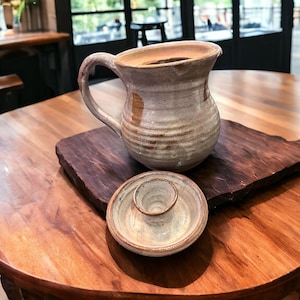 May include: A handmade ceramic pitcher with a handle and a small matching dish sit on a dark wooden serving board. The pitcher is light brown with darker brown accents. The objects are on a round wooden table.