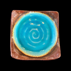 May include: Square ceramic plate with a turquoise spiral design in the center. The plate has a brown rim with an orange accent. The plate is on a black background.