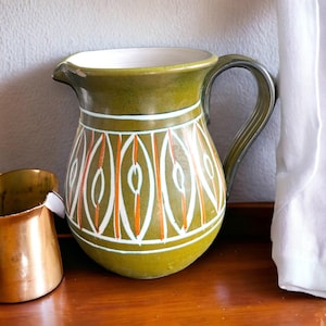 Può includere: Una brocca in ceramica con smalto verde oliva opaco, caratterizzata da un motivo ripetitivo di forme verticali simili a foglie bianche e arancioni sul corpo, abbinata a una piccola tazza in metallo color rame lucido con manico.