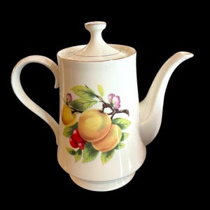 Puede incluir: Tetera de cerámica blanca con tapa y asa. La tetera presenta un colorido diseño de frutas y flores con melocotones, cerezas y hojas verdes. El borde y el asa tienen un ribete dorado. La tetera está sobre un fondo negro.