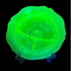 May include: A round, translucent, neon green glass divided plate featuring a central circular medallion and a decorative scalloped rim with an embossed geometric pattern. The glass glows with a bright, vivid luminescence against a black background.
