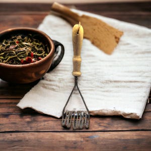 May include: A vintage potato masher with a wooden handle and wire head, resting on a wooden surface. A wooden bowl filled with herbs and spices and a wooden spatula are also visible. The scene evokes a rustic, home-cooked feel.