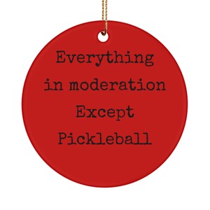 Może przedstawiać: Czerwona, okrągła ozdoba choinkowa z napisem "Wszystko z umiarem, oprócz pickleballa" w kolorze czarnym.