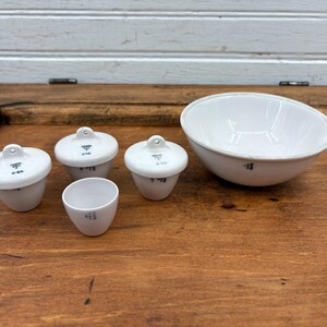 May include: Set of five white ceramic laboratory crucibles. The crucibles are different sizes and have black markings. The largest crucible is a bowl shape.