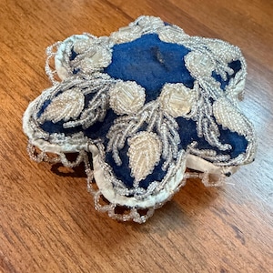 May include: A star-shaped, vintage-style pin cushion with a deep blue center and white trim. The cushion is embellished with silver-colored beads and leaf-shaped accents, creating a decorative and ornate design.