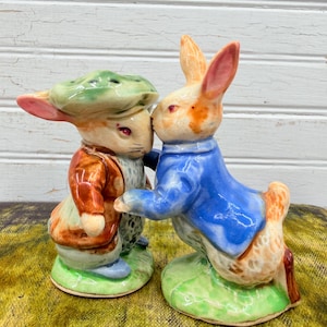 May include: A pair of ceramic rabbit figurines, one in a blue jacket and the other in a brown jacket and green hat, are depicted nose-to-nose. The figurines are on green bases, with a white and light brown color scheme.