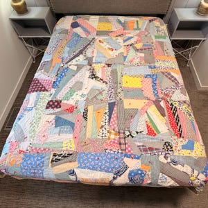 May include: A patchwork quilt featuring a chaotic arrangement of various fabric scraps in diverse colors, patterns, and textures. The quilt is constructed from irregular geometric shapes stitched together, accented with small red yarn ties.