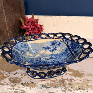May include: A blue and white ceramic serving dish with a landscape design. The oval-shaped dish has a pedestal base and a decorative, openwork rim. The interior features a detailed scene of trees and a building.