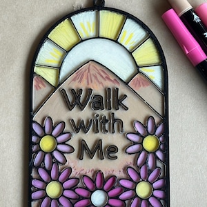 Puede incluir: Placa decorativa arqueada con las palabras "Walk with Me". El dise&ntilde;o presenta un sol amarillo, monta&ntilde;as y margaritas moradas. La placa est&aacute; enmarcada en negro.