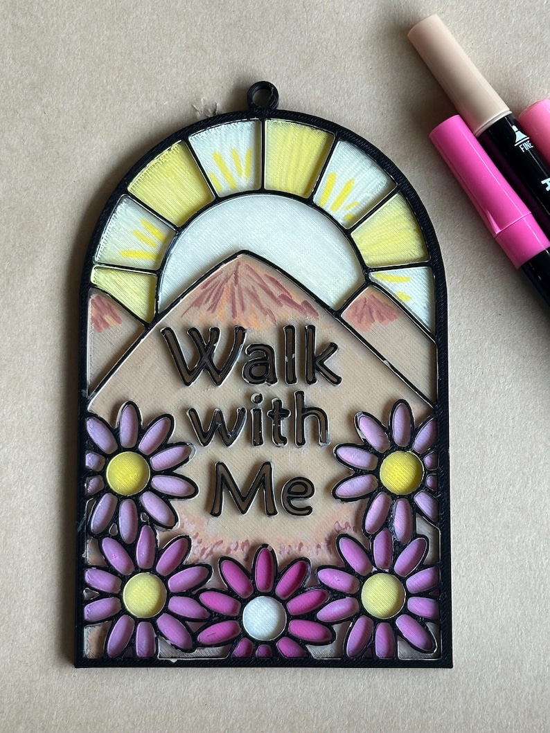 Puede incluir: Placa decorativa arqueada con las palabras "Walk with Me". El dise&ntilde;o presenta un sol amarillo, monta&ntilde;as y margaritas moradas. La placa est&aacute; enmarcada en negro.