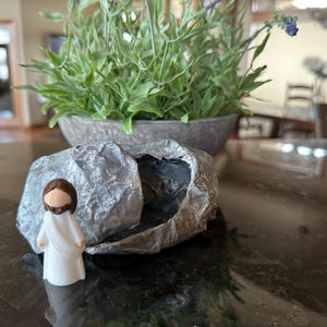 May include: A small white figurine of Jesus standing outside of a gray rock tomb with the top half of the rock rolled away. The tomb is on a dark brown surface.