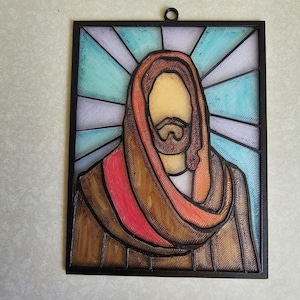 Puede incluir: Una obra de arte enmarcada al estilo vidriera que representa una figura religiosa. La figura tiene barba marr&oacute;n y viste una t&uacute;nica marr&oacute;n y roja. El fondo presenta un patr&oacute;n radiante azul y blanco, todo dentro de un marco negro.