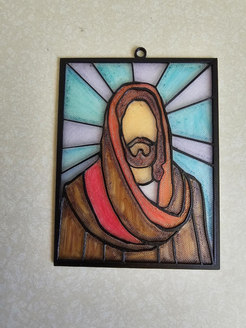 Puede incluir: Una obra de arte enmarcada al estilo vidriera que representa una figura religiosa. La figura tiene barba marr&oacute;n y viste una t&uacute;nica marr&oacute;n y roja. El fondo presenta un patr&oacute;n radiante azul y blanco, todo dentro de un marco negro.