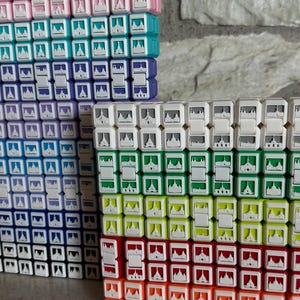 May include: A collection of colorful, interlocking cube toys. The cubes are arranged in stacks and feature a repeating design of a building. Colors include pink, teal, purple, blue, white, green, yellow, red, and orange.