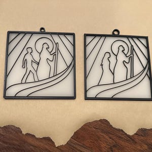 Puede incluir: Dos adornos cuadrados con marco negro e interiores blancos. Cada uno presenta una silueta de figuras, una con dos personas y la otra con un par. Los dise&ntilde;os recuerdan a las vidrieras, con un lazo en la parte superior para colgar.