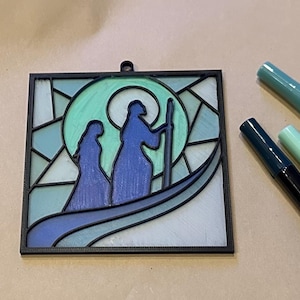 Puede incluir: Un adorno de pared estilo vidriera que representa una escena religiosa con dos figuras azules y un halo verde. La obra de arte est&aacute; enmarcada en negro y tiene un lazo para colgar. Tres rotuladores verdes y azules en el fondo.