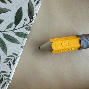 May include: A yellow pencil-shaped object with the word "Choose" and "The Right" printed on it. The pencil has a gray band and a pink eraser. A notebook with a green and white leaf pattern is in the background.