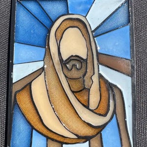 Puede incluir: Panel de vidriera que representa una figura religiosa con barba y t&uacute;nica. La obra de arte presenta una paleta de colores azul, marr&oacute;n y beige, con un borde negro. El dise&ntilde;o recuerda a las vidrieras de las iglesias.