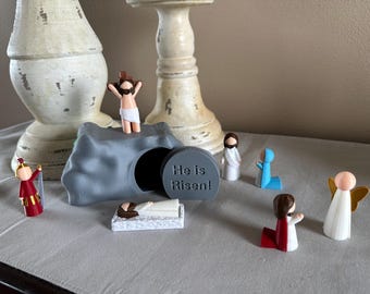 May include: A gray stone tomb with a lid that reads "He is Risen!"  A small figurine of Jesus is standing on top of the tomb.  There are several other figurines around the tomb, including a kneeling angel, a soldier, and a woman holding a white cloth.