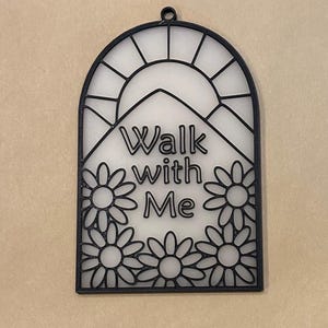 Puede incluir: Adorno de pared negro en forma de arco con el texto "Walk with Me" y un dise&ntilde;o floral. El dise&ntilde;o incluye un sol y flores tipo margarita. El adorno de pared tiene un peque&ntilde;o lazo en la parte superior para colgar.