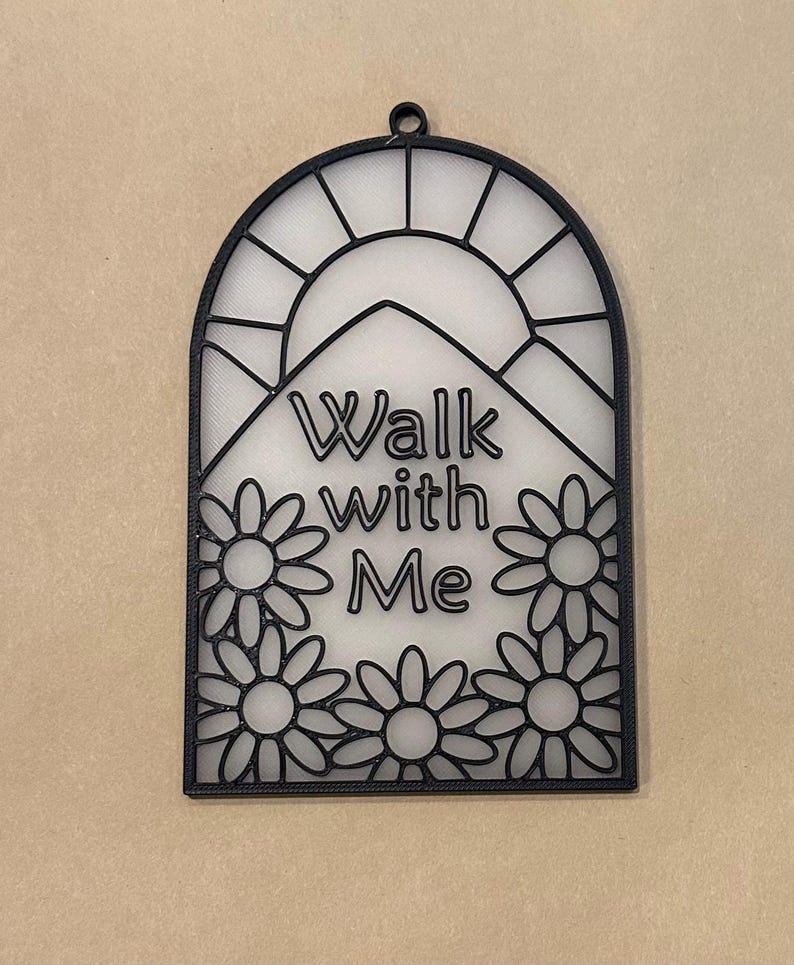 Puede incluir: Adorno de pared negro en forma de arco con el texto "Walk with Me" y un dise&ntilde;o floral. El dise&ntilde;o incluye un sol y flores tipo margarita. El adorno de pared tiene un peque&ntilde;o lazo en la parte superior para colgar.