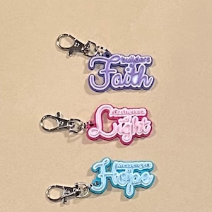 May include: Three keychains with silver clasps. The first keychain is purple and white and reads "Builders of Faith." The second keychain is pink and white and reads "Gatherers of Light." The third keychain is blue and white and reads "Messengers of Hope."