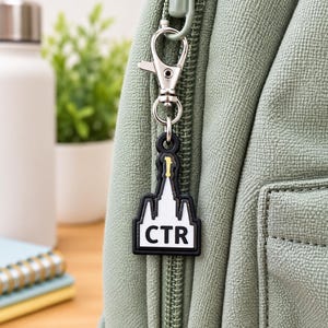 May include: A black and white rubber keychain shaped like a temple spire with the letters CTR in bold black font. The charm features a small yellow accent at the top and is attached to a silver-colored metal lobster clasp hook on a sage green bag.