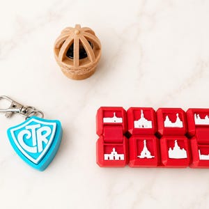 May include: A tan 3D-printed dome-shaped object, a light blue shield-shaped keychain with the letters CTR in white, and a red rectangular folding fidget toy featuring eight white silhouettes of various building structures on its surface.