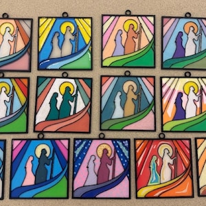 Puede incluir: Una colecci&oacute;n de doce colgantes de vidrieras. Cada panel cuadrado presenta una escena religiosa con figuras en silueta, sobre un fondo de colores vibrantes, incluyendo azul, verde y naranja. Los paneles tienen un borde negro.