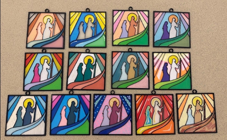 Puede incluir: Una colecci&oacute;n de doce colgantes de vidrieras. Cada panel cuadrado presenta una escena religiosa con figuras en silueta, sobre un fondo de colores vibrantes, incluyendo azul, verde y naranja. Los paneles tienen un borde negro.